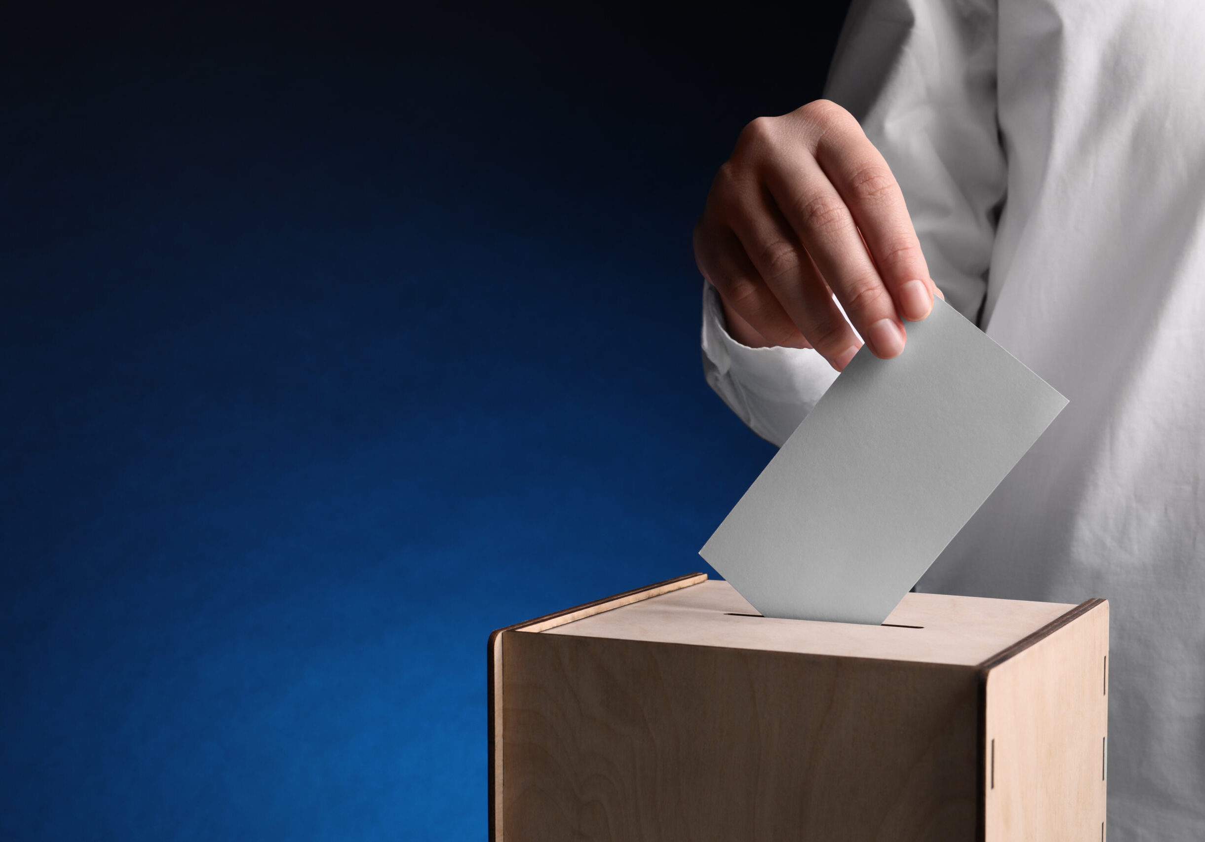 Woman putting her vote into ballot box on dark blue background, closeup. Space for text